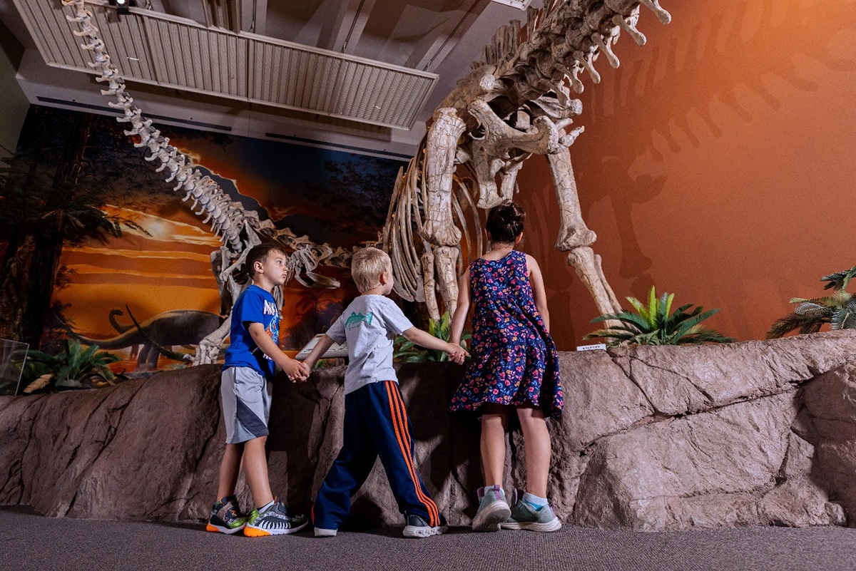 Three children hold hands while viewing a large dinosaur skeleton